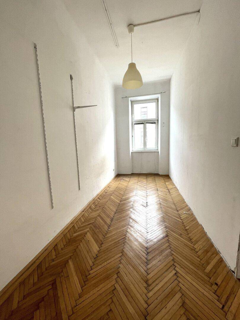 Schmaler Raum mit Fischgrätparkett und einem Fenster, das Tageslicht hereinlässt.