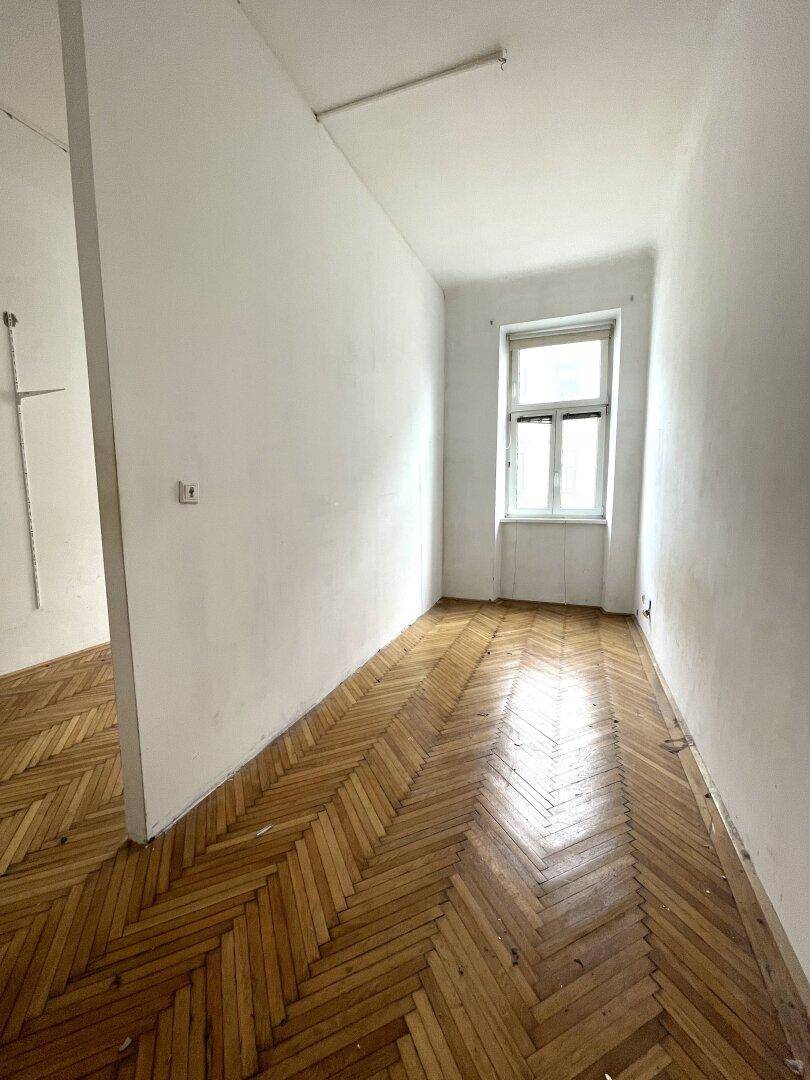 Heller Raum mit Fischgrätparkett, Fenster und einer Trennwand, die den Bereich strukturiert.