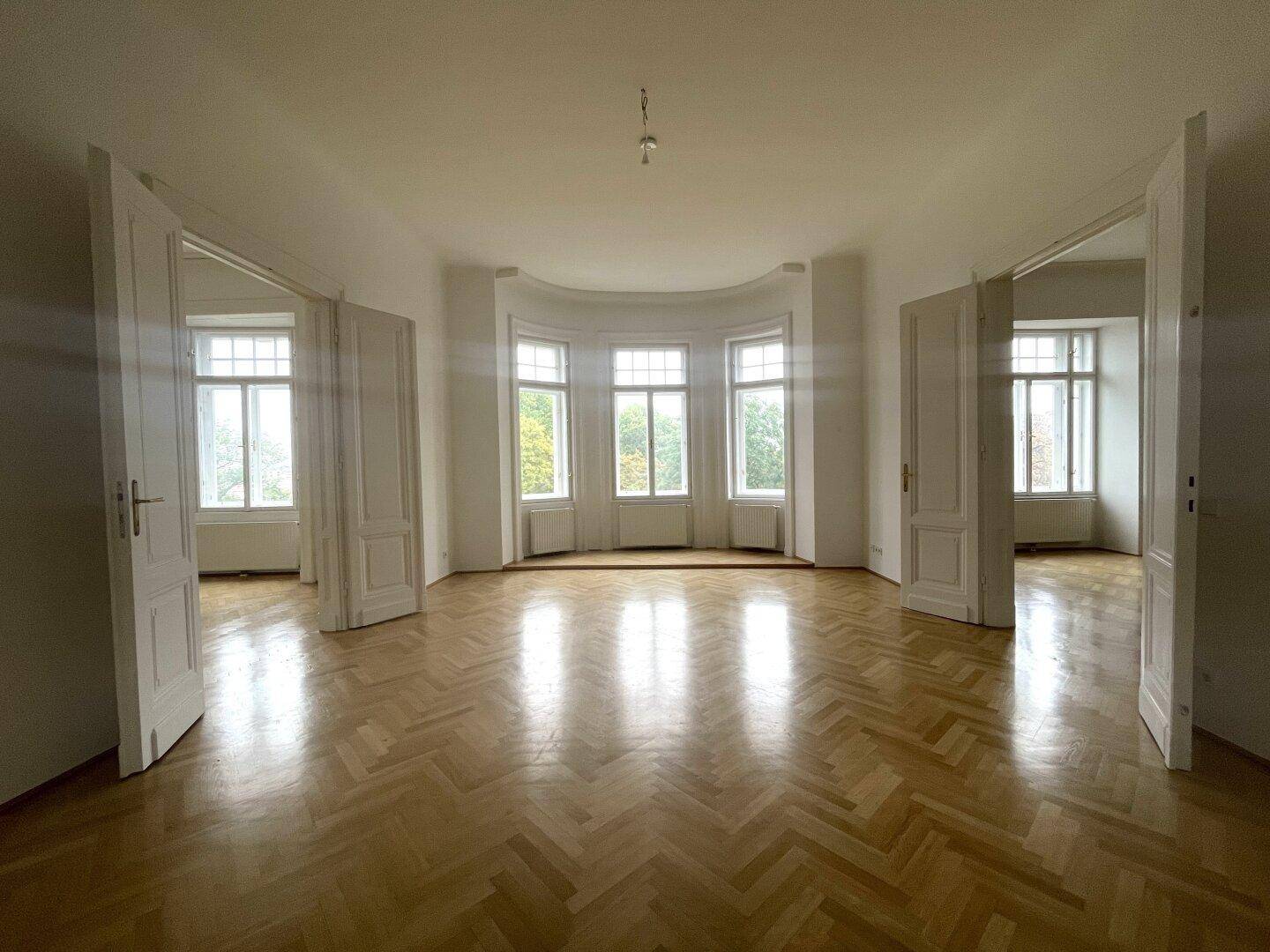 Großes, leeres Zimmer mit Fischgrätparkett, hohen Decken und einem Erker mit vielen Fenstern.