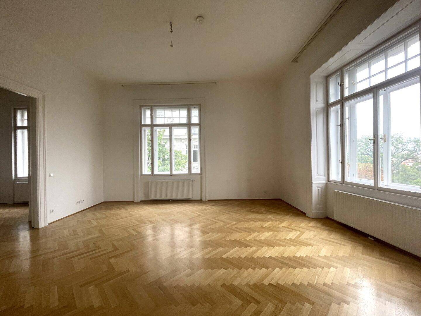 Geräumiges Zimmer mit klassischem Fischgrätparkett, hohen Fenstern und viel Tageslicht.