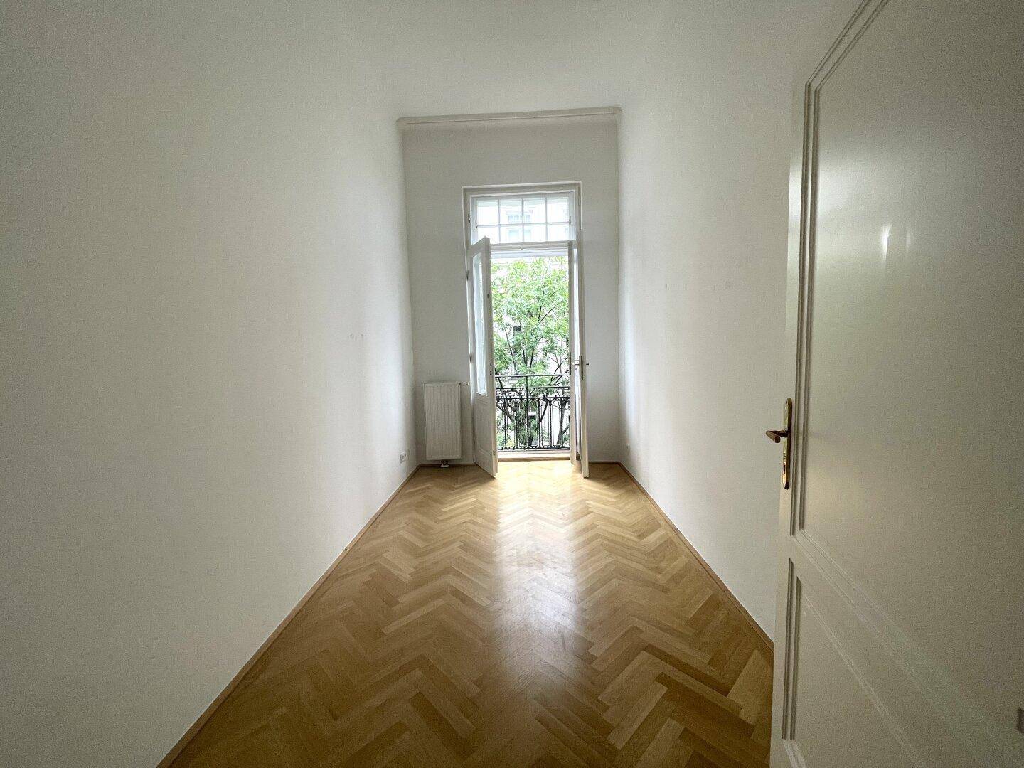 Schmaler Raum mit Fischgrätparkett und Zugang zu einem französischen Balkon mit Blick ins Grüne.