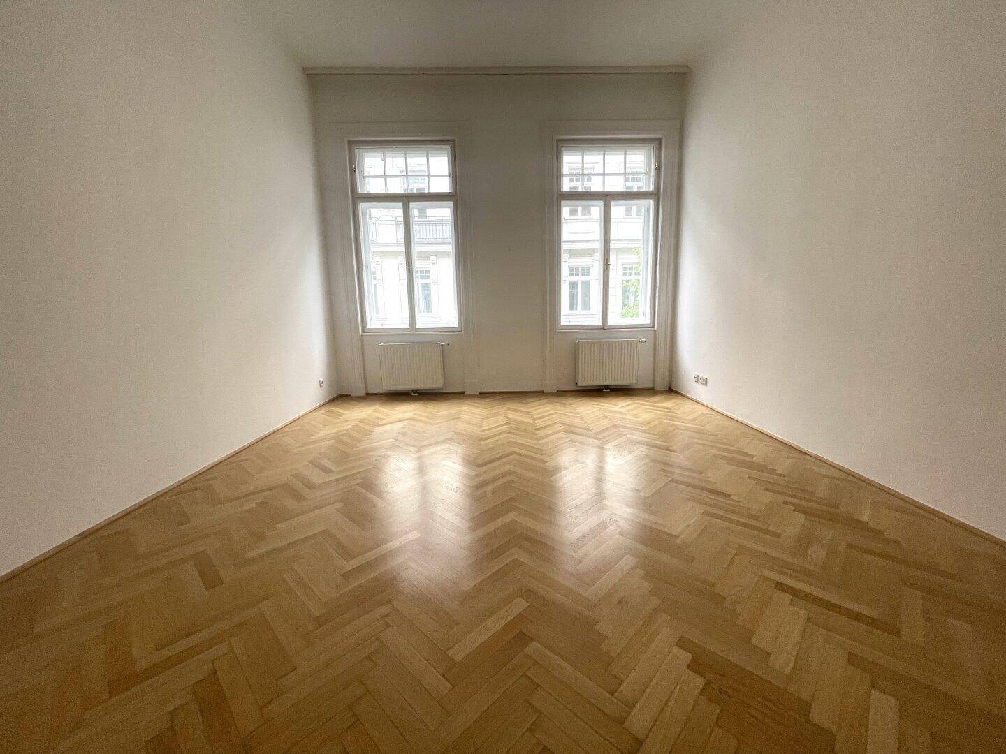 Leeres Zimmer mit Fischgrätparkett und zwei großen Fenstern mit Blick auf die gegenüberliegende Bebauung.