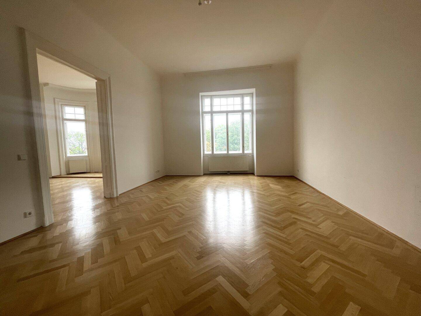 Helles Zimmer mit elegantem Fischgrätparkett und einem großen Fenster, das viel Licht hereinlässt.