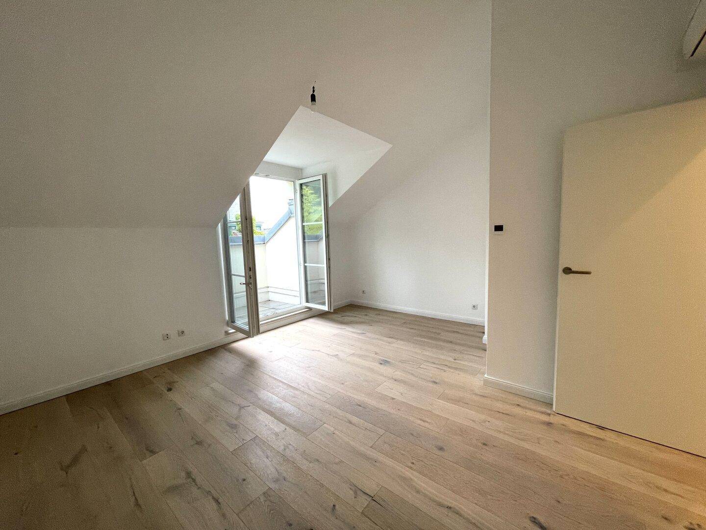 Geräumiges Schlafzimmer mit hellem Parkettboden, Dachschrägen und Zugang zu einem kleinen Balkon.