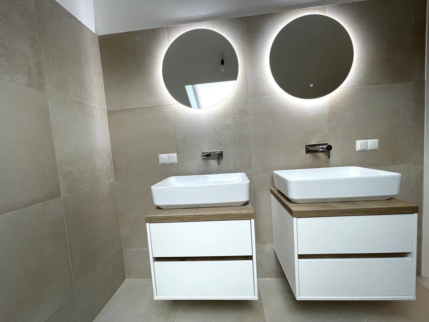Modernes Badezimmer mit Doppelwaschbecken, beleuchteten Spiegeln und großformatigen Fliesen.