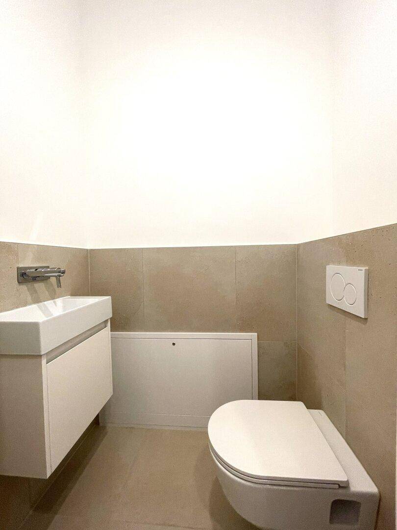 Modernes Gäste-WC mit wandhängender Toilette, kleinem Waschbecken und beigen Fliesen.