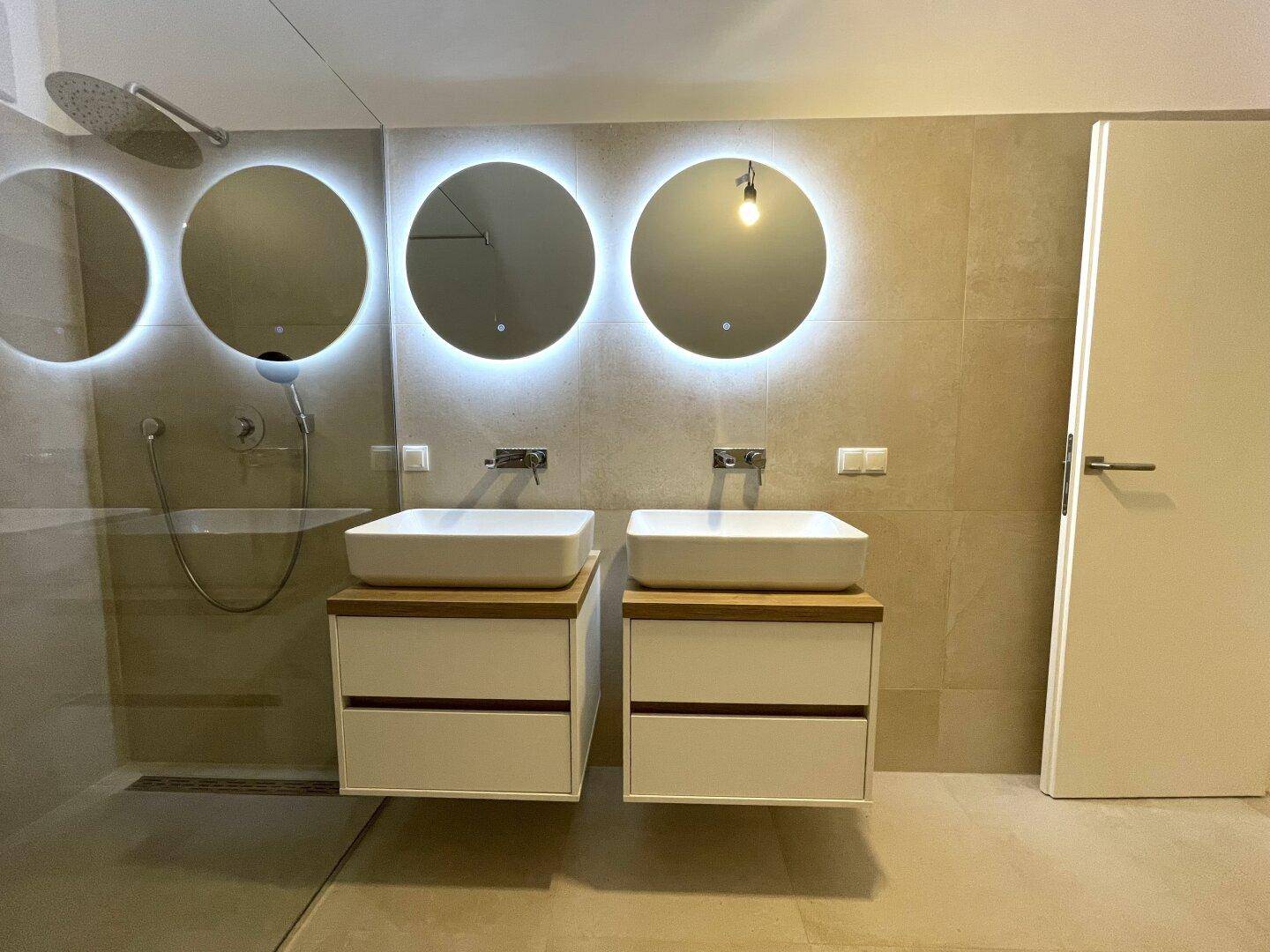 Modernes Badezimmer mit begehbarer Dusche, Doppelwaschbecken und beleuchteten Spiegeln.