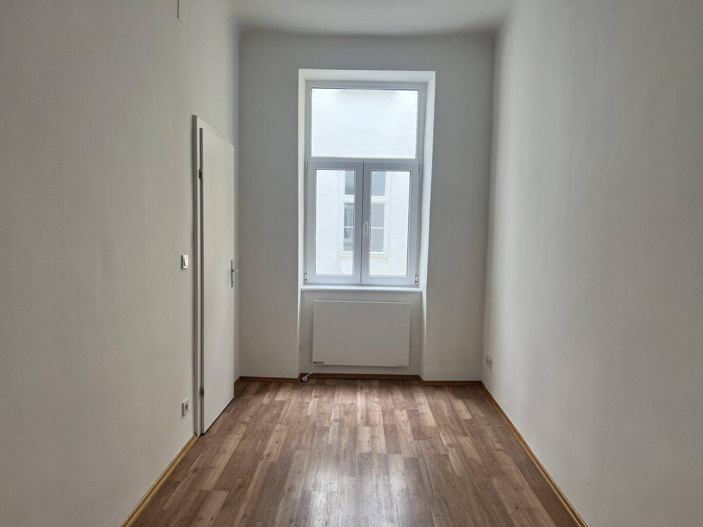 Schmaler Raum mit Laminatboden, einem großen Fenster und weiß gestrichenen Wänden, ideal als Schlafzimmer.