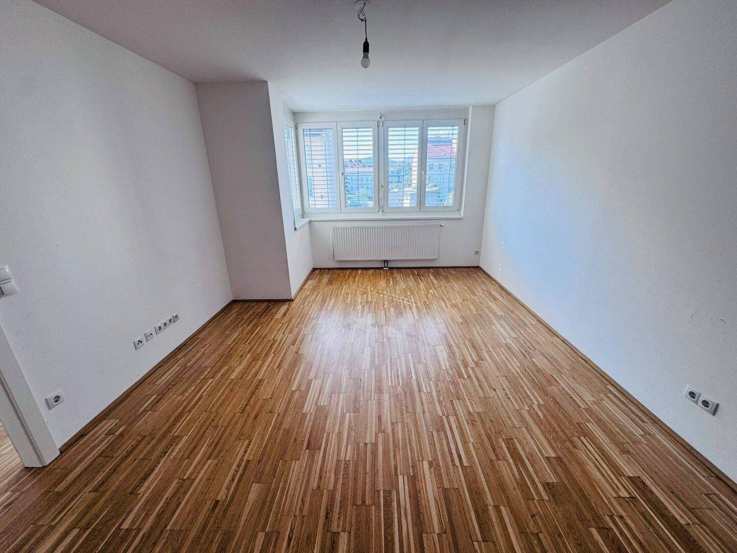 Helles Schlafzimmer mit Parkettboden, weißen Wänden und einem Fenster mit Blick nach draußen.