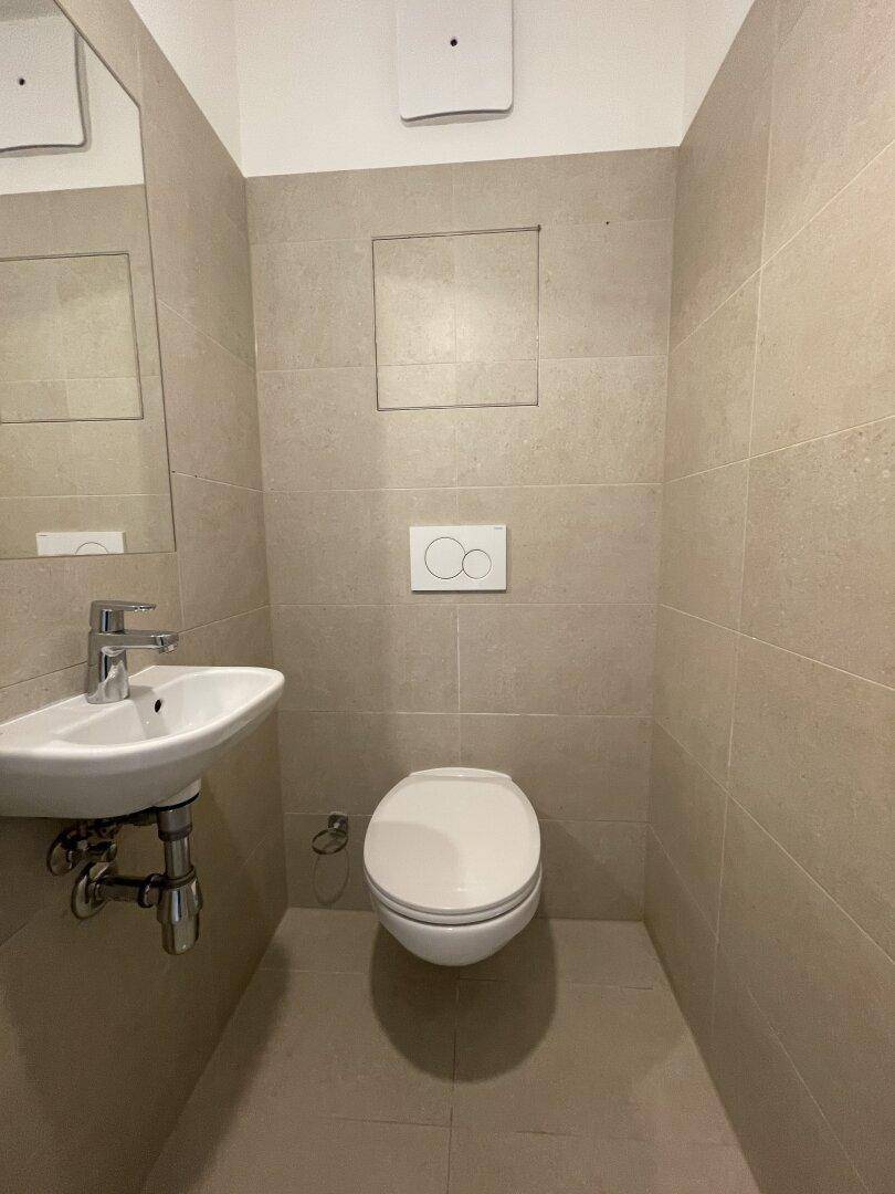 Separates WC mit wandhängender Toilette und kleinem Waschbecken, komplett gefliest in Beige.
