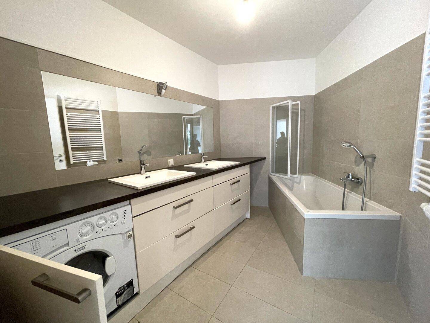 Modernes Badezimmer mit Doppelwaschbecken, Badewanne und integrierter Waschmaschine, hell gefliest.