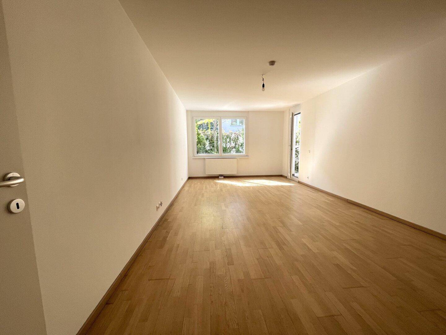 Heller, leerer Wohnraum mit hellem Parkettboden und großen Fenstern, die viel Tageslicht hereinlassen.