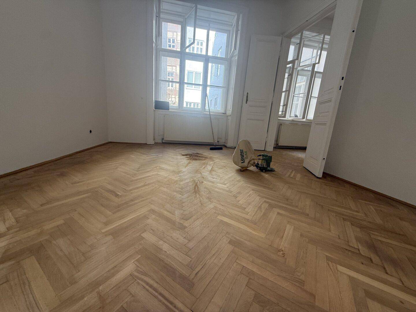 Leeres Zimmer mit Fischgrätparkett, großen Fenstern und Blick auf die Stadt.