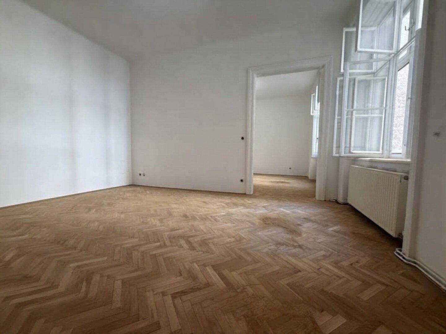 Geräumiges, leeres Zimmer mit Fischgrätparkett, hohen Decken und großen Fenstern.