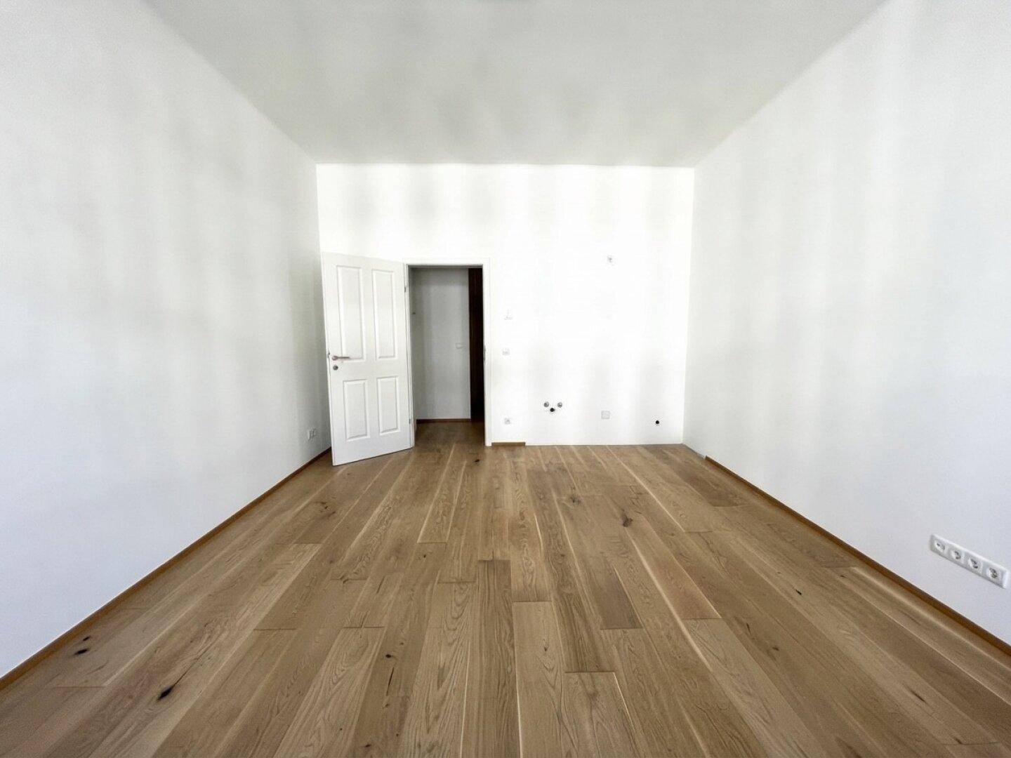 Geräumiges, leeres Zimmer mit Holzboden und weißer Tür, ideal für verschiedene Nutzungen.