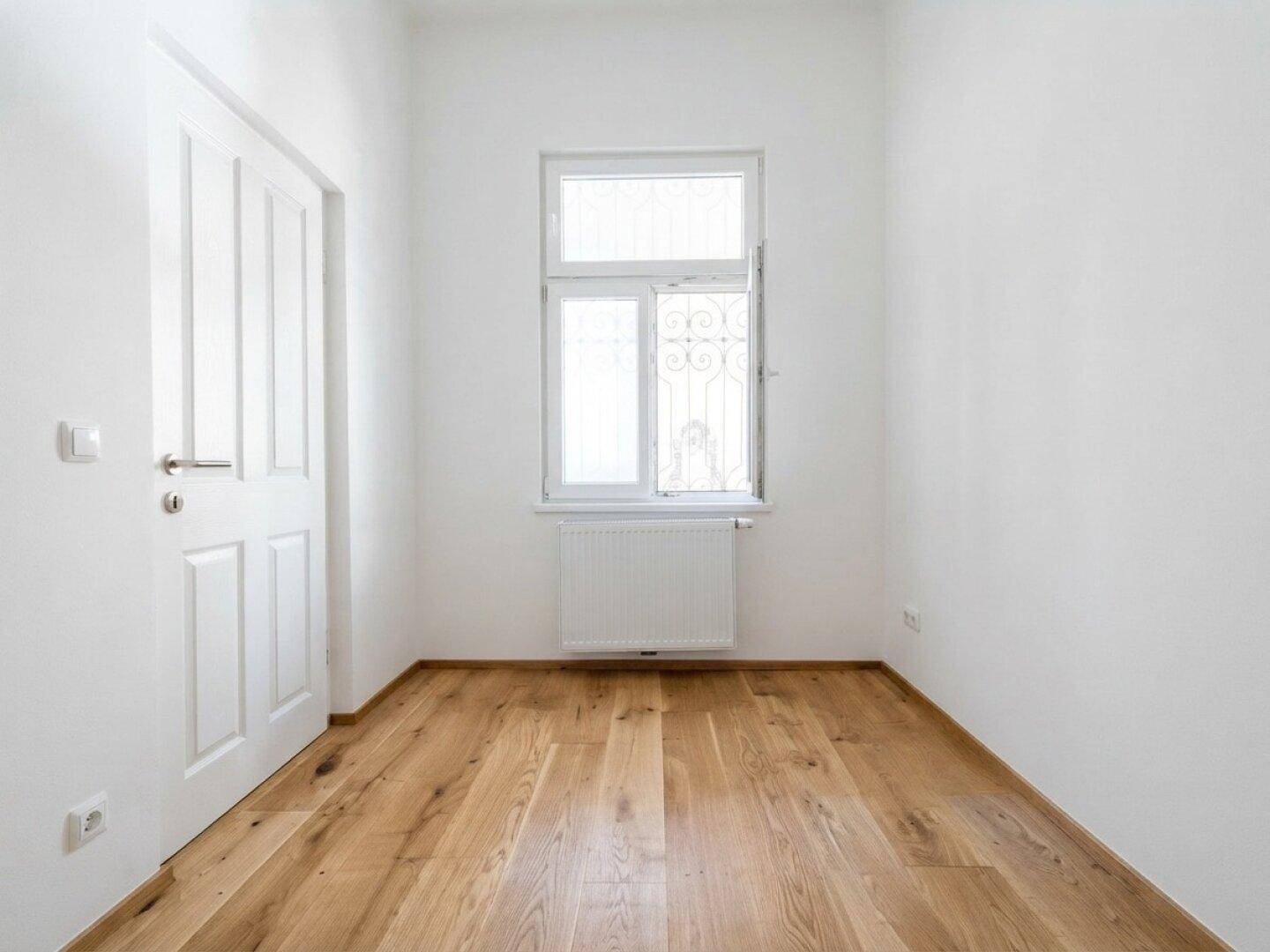 Leeres Zimmer mit Holzboden, einem Fenster und Heizkörper, bereit für neue Einrichtung.