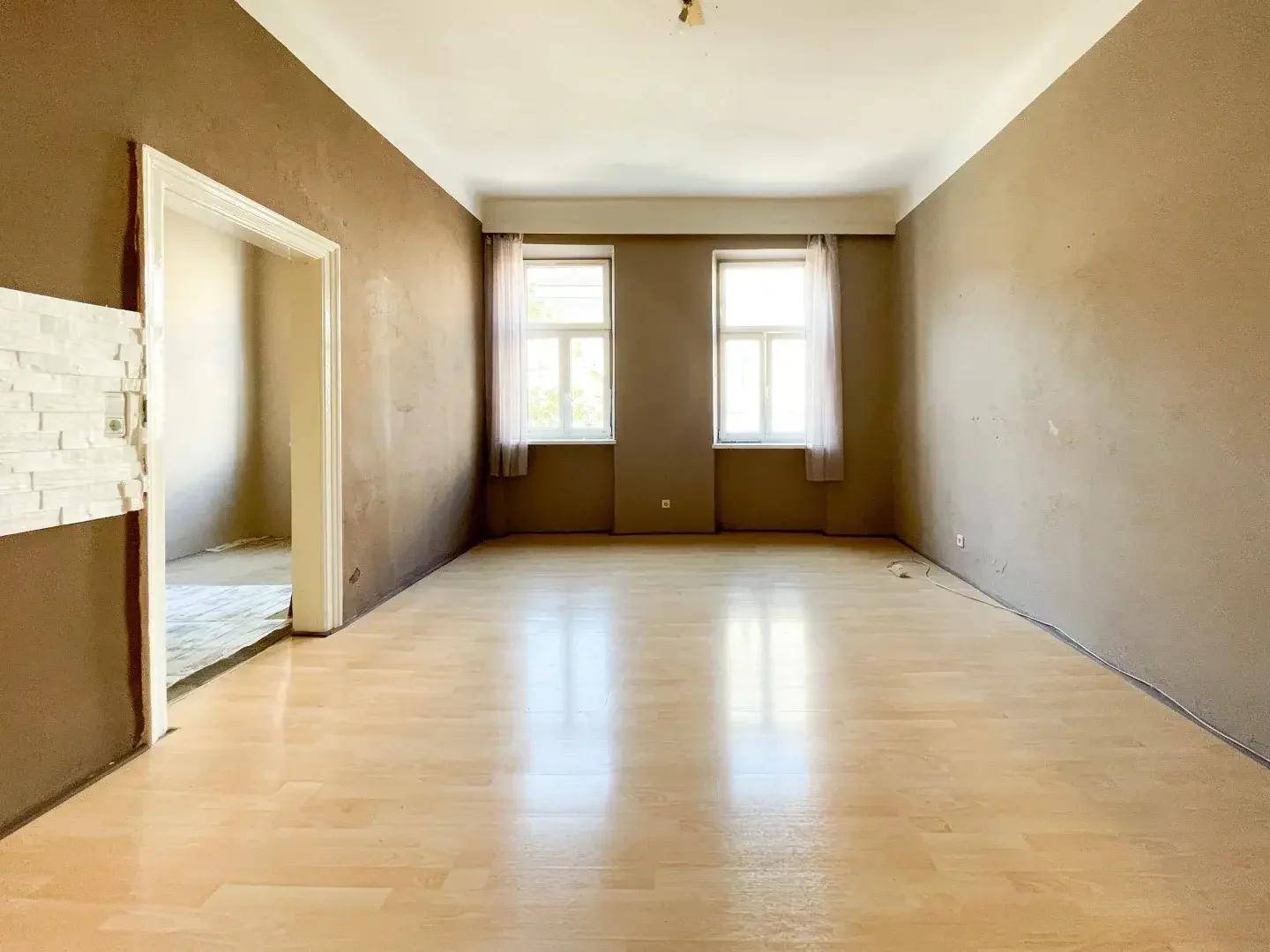 Geräumiges Wohnzimmer mit hellem Laminatboden, zwei Fenstern und Wänden in warmen Erdtönen.