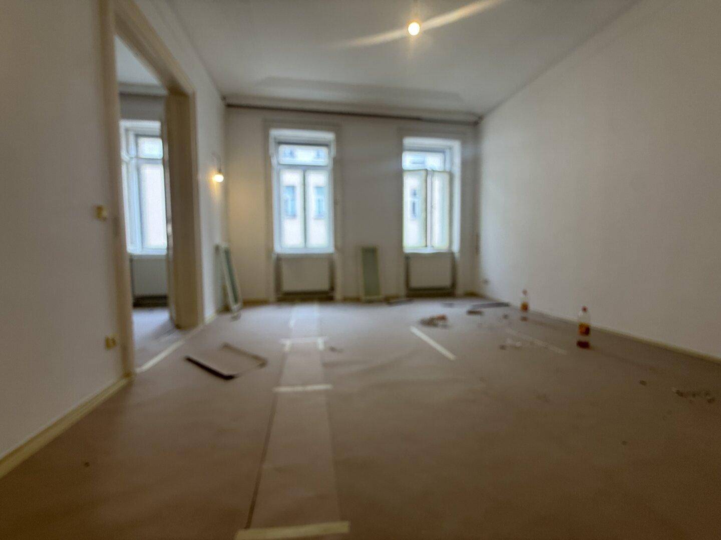 Heller Raum mit Renovierungsarbeiten, bedecktem Boden und Blick durch zwei Fenster.