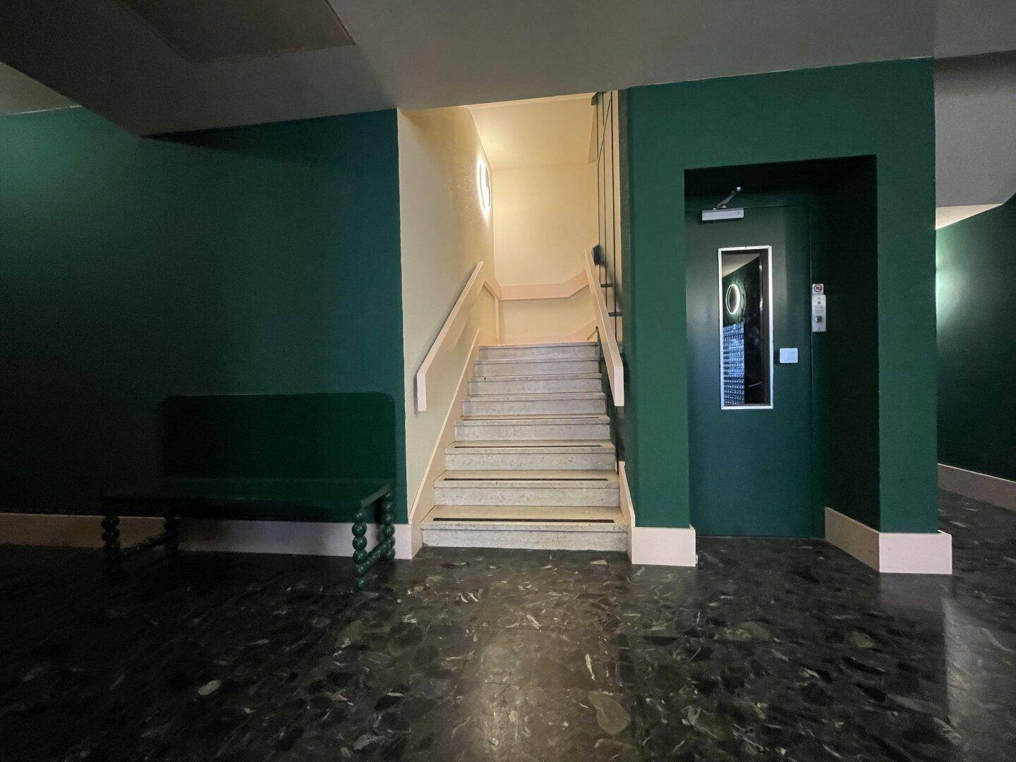 Eingangsbereich mit dunklem Fliesenboden, grünen Wänden, Treppe und Aufzugzugang.