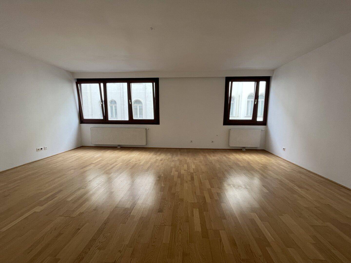 Geräumiges Wohnzimmer mit hellem Parkettboden und zwei großen Fenstern für viel Tageslicht.