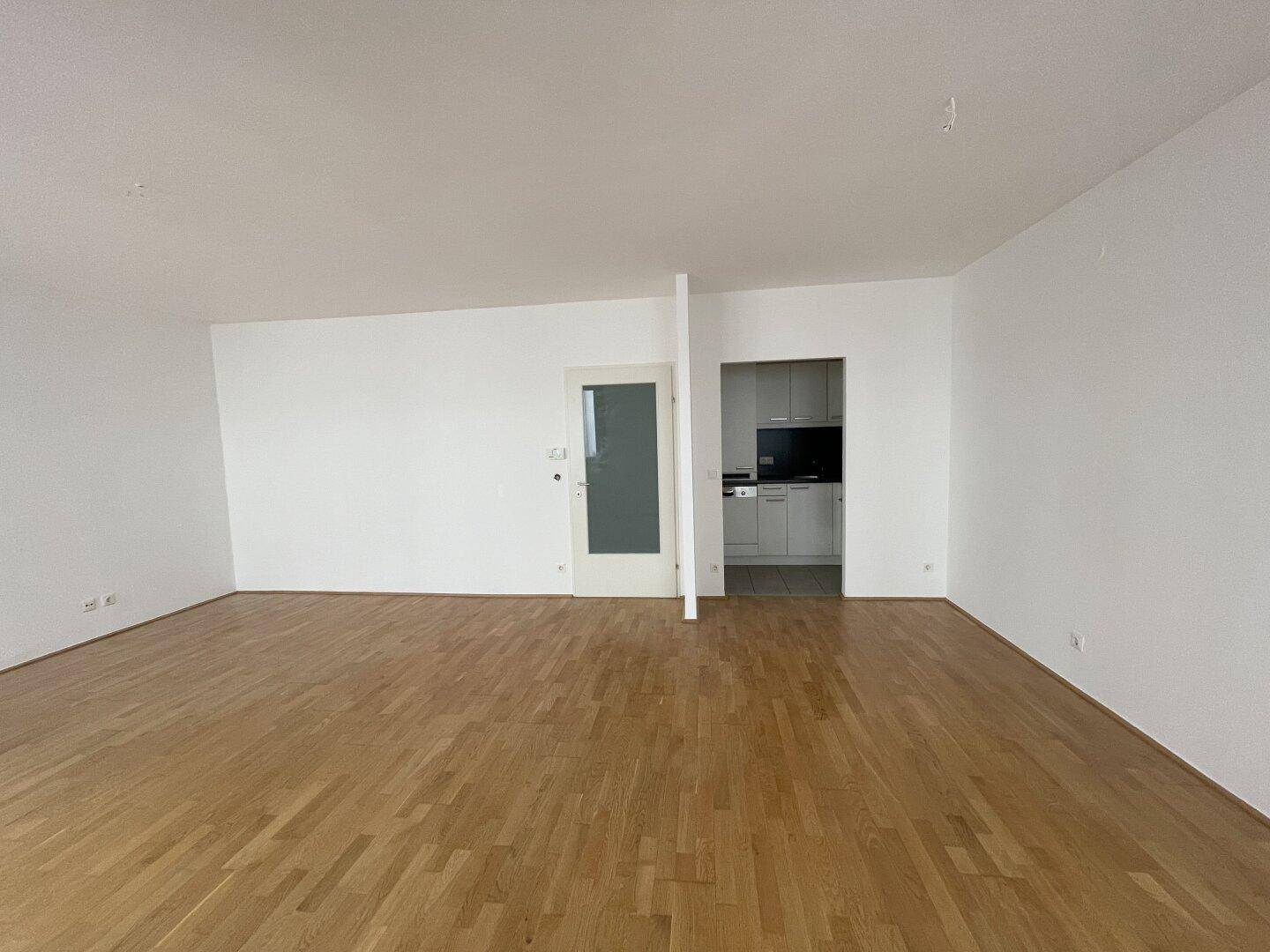 Geräumiges Wohnzimmer mit Parkettboden und Zugang zu einem weiteren Raum, möglicherweise der Küche.
