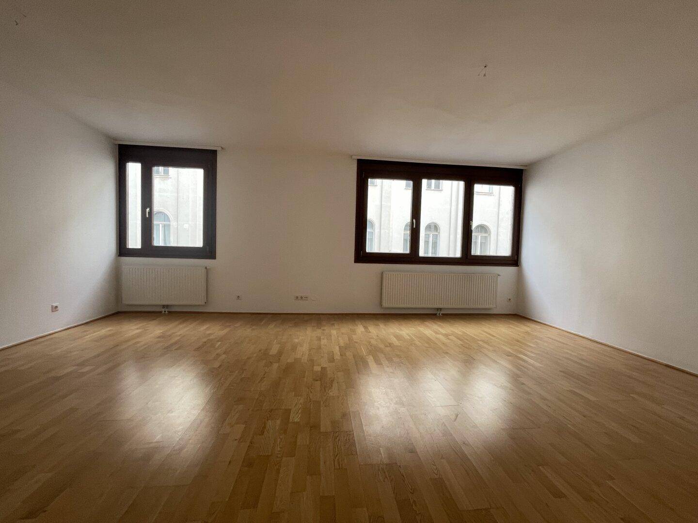 Leeres Wohnzimmer mit hellen Wänden, Parkettboden und zwei großen Fenstern, die den Raum erhellen.