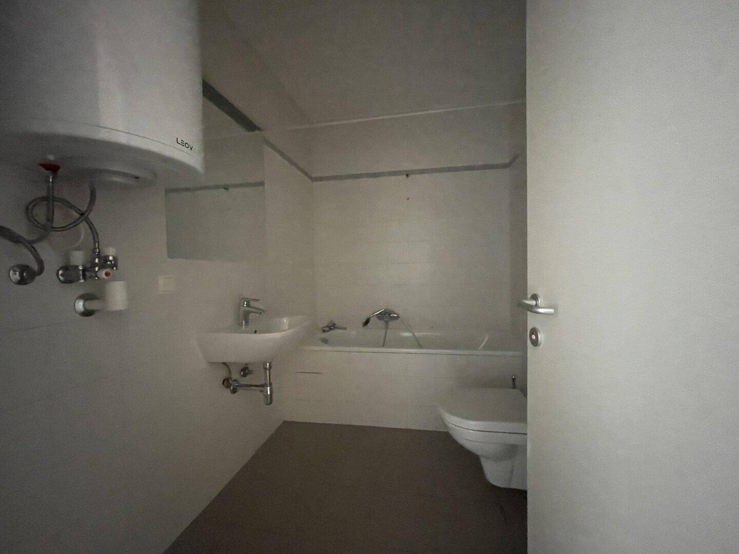 Schlichtes Badezimmer mit Badewanne, Waschbecken, Toilette und einem Warmwasserboiler an der Wand.