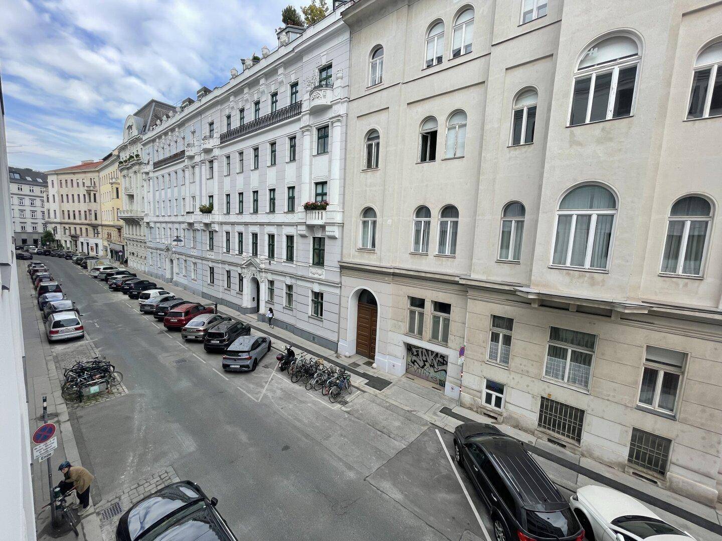 Blick von oben auf eine belebte Stadtstraße mit parkenden Autos und historischen Gebäudefassaden.