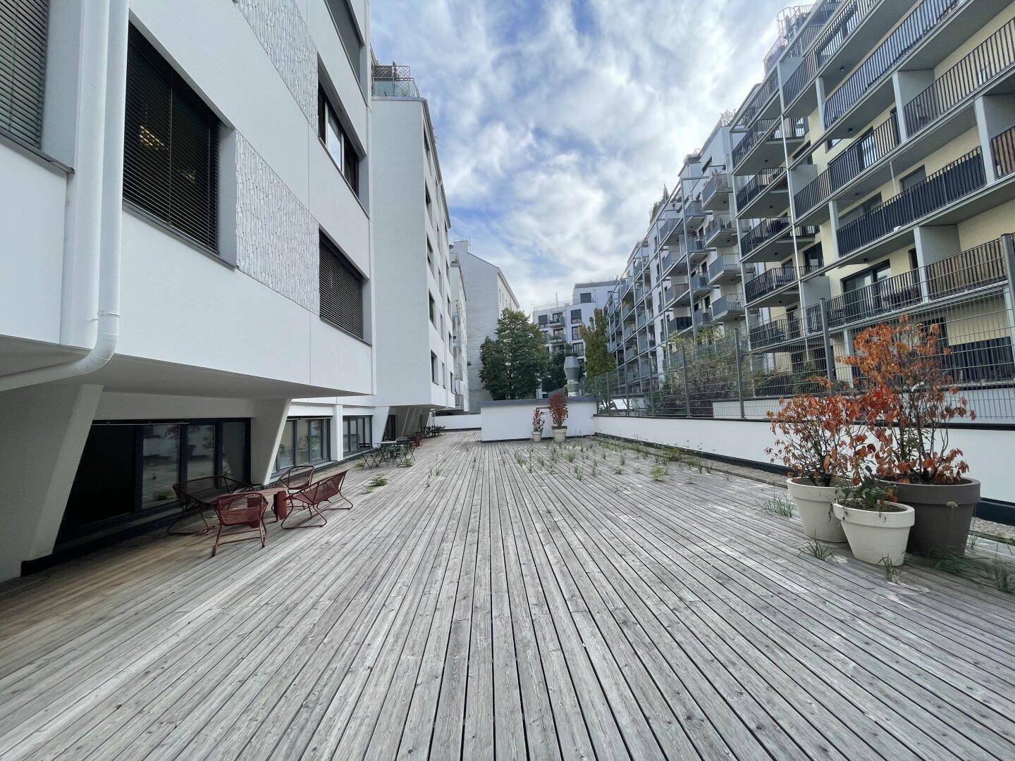 Große Holzterrasse im Freien, umgeben von modernen Wohngebäuden mit Balkonen.