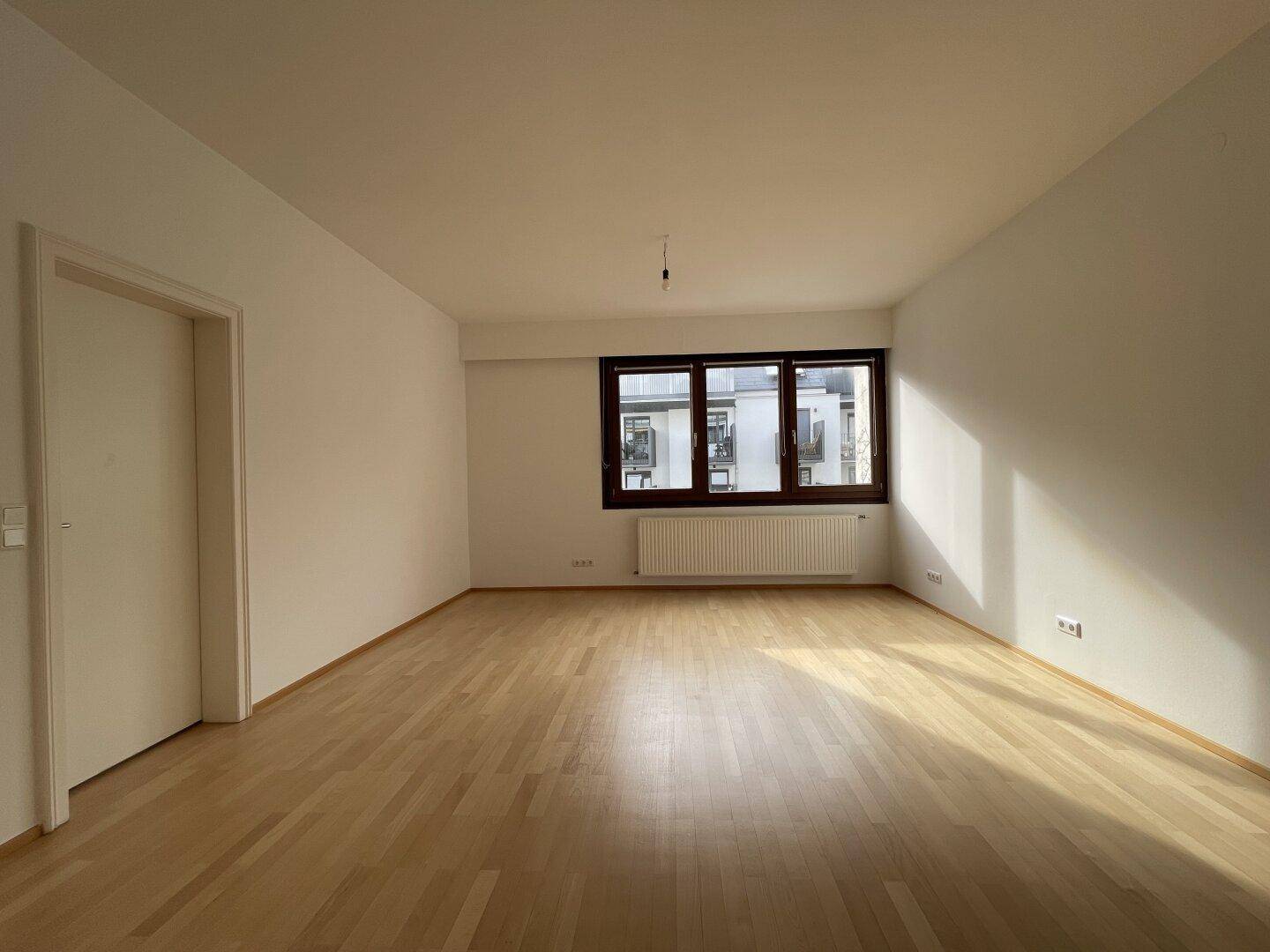 Geräumiges, leeres Wohnzimmer mit hellem Parkettboden und einem großen Fenster.