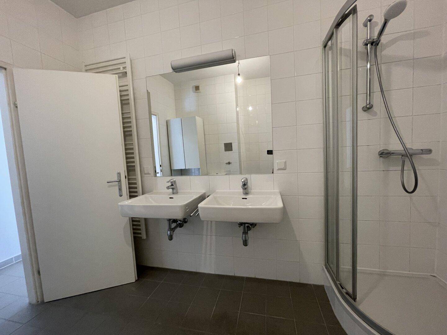 Helles Badezimmer mit Doppelwaschbecken, Dusche und großem Spiegel, weiß gefliest.