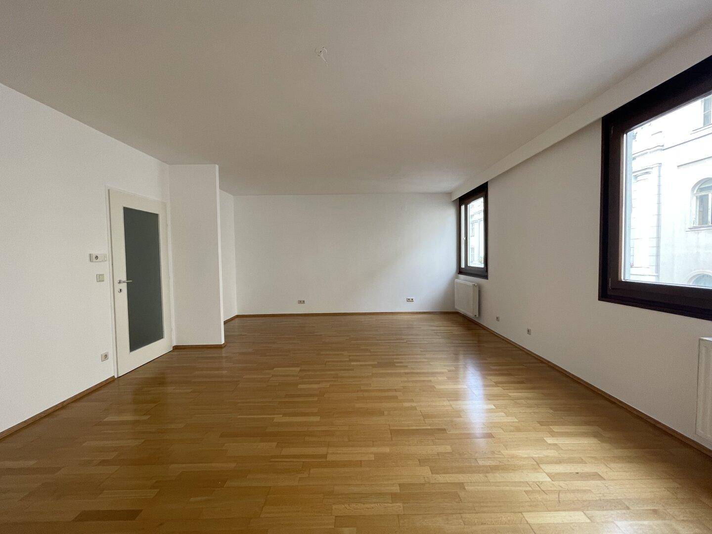 Geräumiges Wohnzimmer mit hellem Parkettboden und viel Tageslicht durch die Fenster.