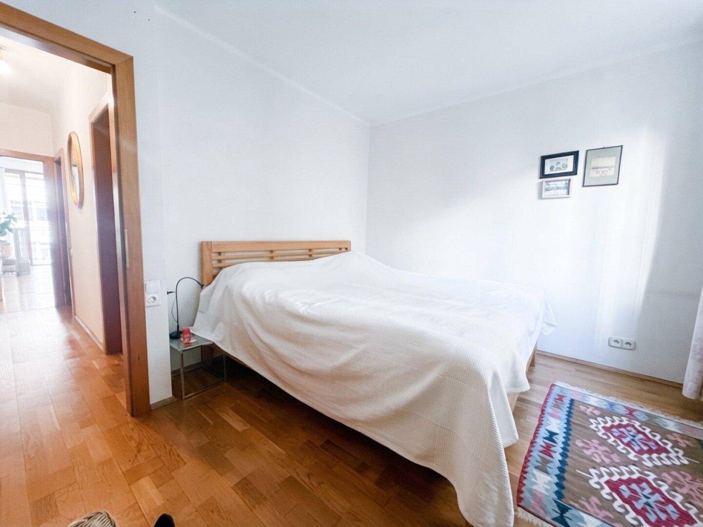 Schlafzimmer mit Holzbett, weißer Bettwäsche und einem farbenfrohen Teppich, mit Zugang zu einem weiteren Raum.