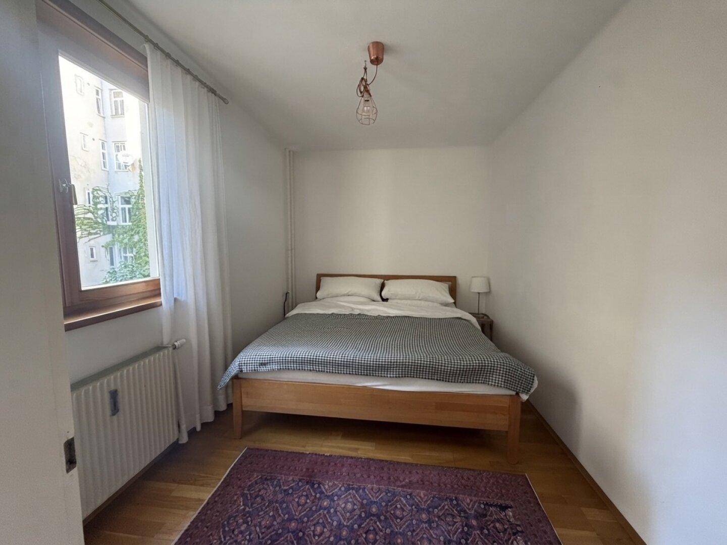 Helles Schlafzimmer mit Holzbett, Fenster und Heizkörper, bietet eine ruhige Atmosphäre.
