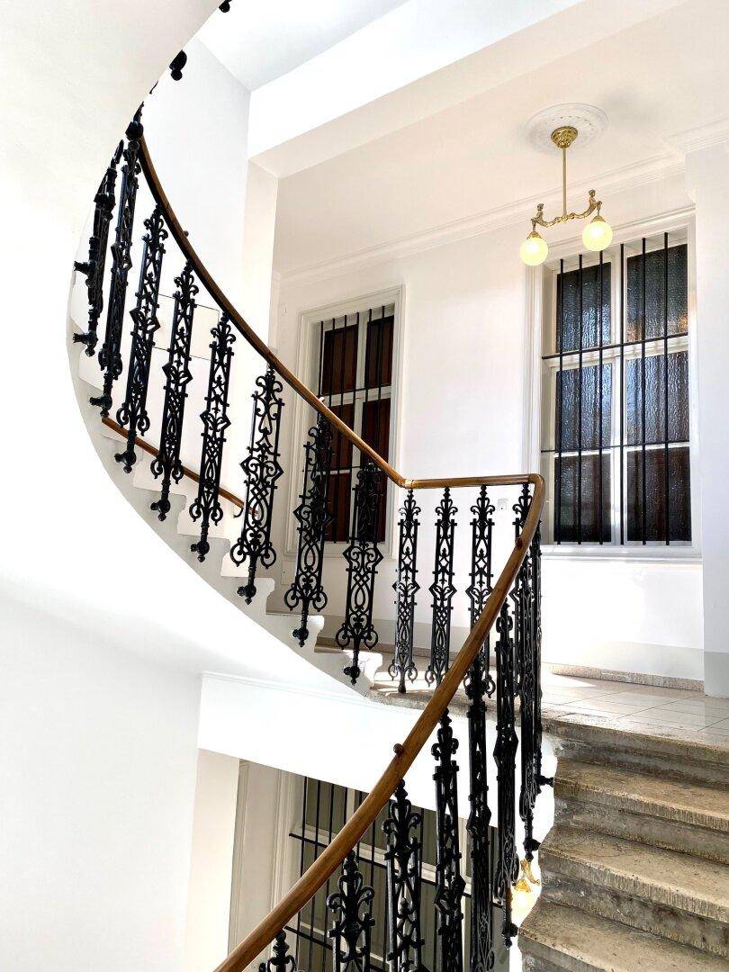 Detailaufnahme einer eleganten Wendeltreppe mit kunstvollem schmiedeeisernem Geländer und großen Fenstern.