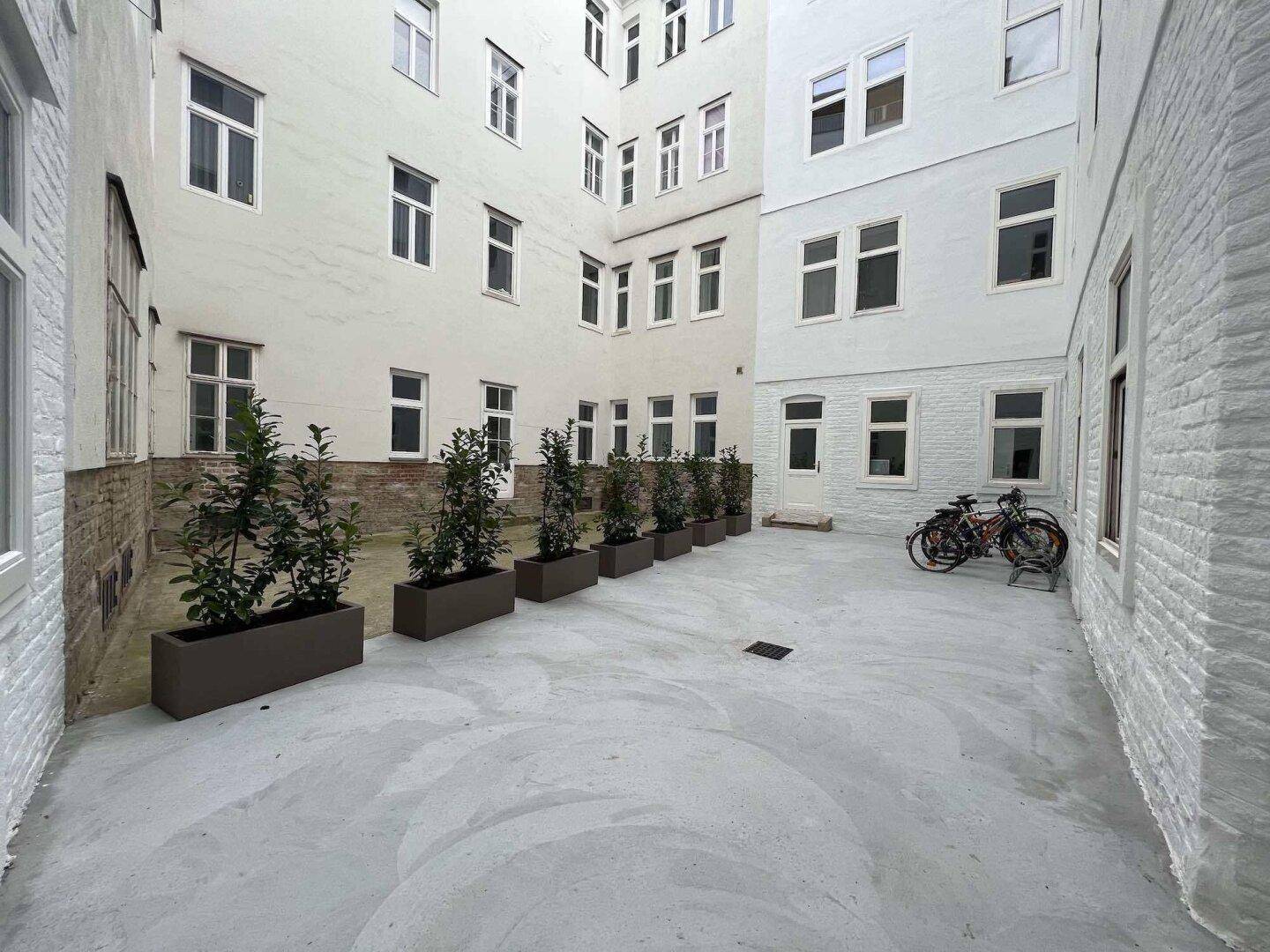 Heller Innenhof mit Pflanzenkübeln und Fahrradstellplätzen, umgeben von Gebäudefassaden.