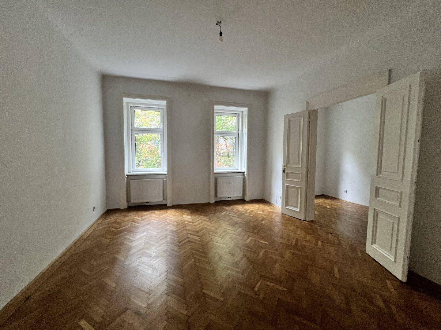 Geräumiges, helles Zimmer mit klassischem Fischgrätparkett und zwei Fenstern, ideal für Wohnzwecke.