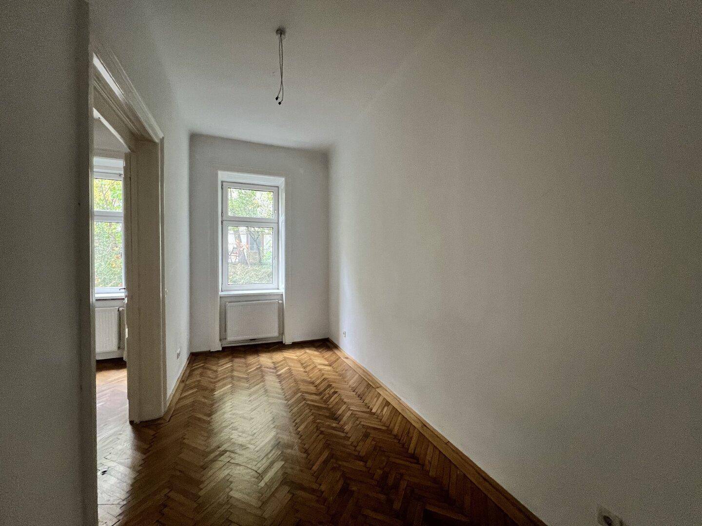Länglicher Raum mit klassischem Fischgrätparkett und Fenster, bietet vielseitige Einrichtungsmöglichkeiten.