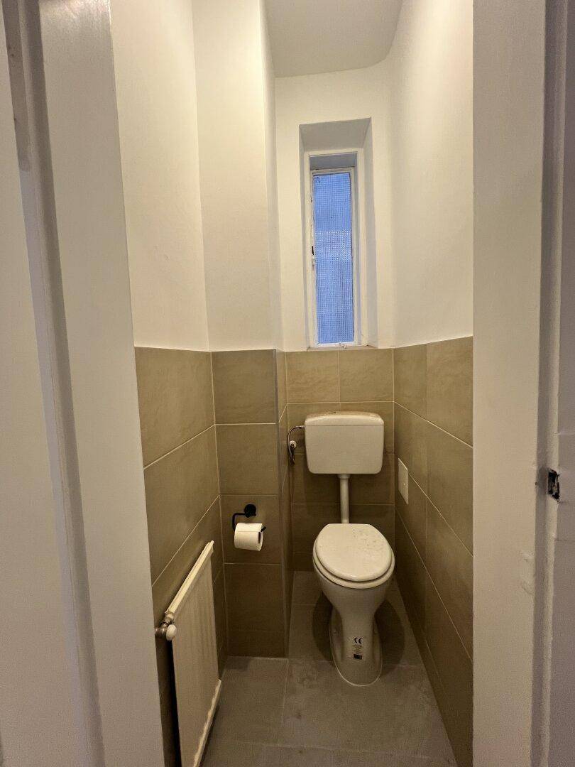 Separate Toilette mit kleinem Fenster und beigen Wandfliesen, funktional und hell.