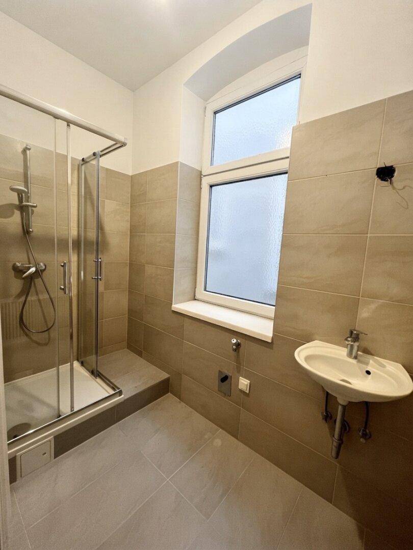 Modernes Badezimmer mit Duschkabine, Waschbecken und Fenster, ausgestattet mit beigen Fliesen.