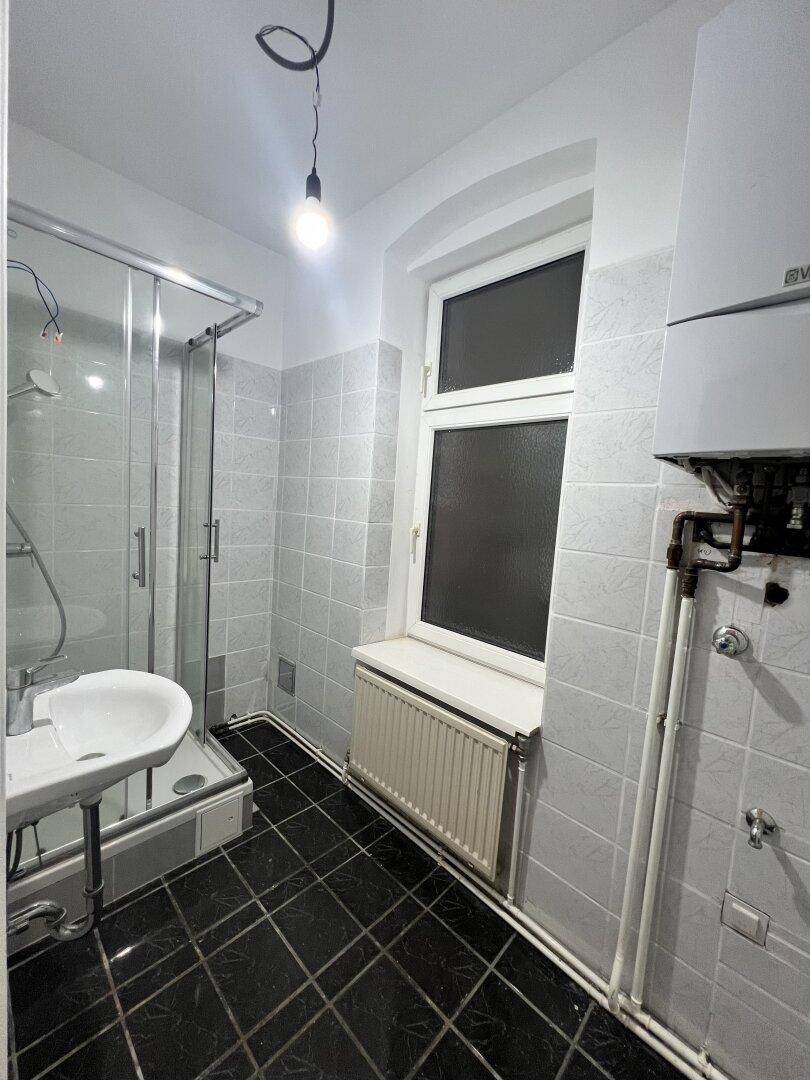 Badezimmer mit Duschkabine, Waschbecken, Fliesenboden und einem Fenster für Tageslicht.