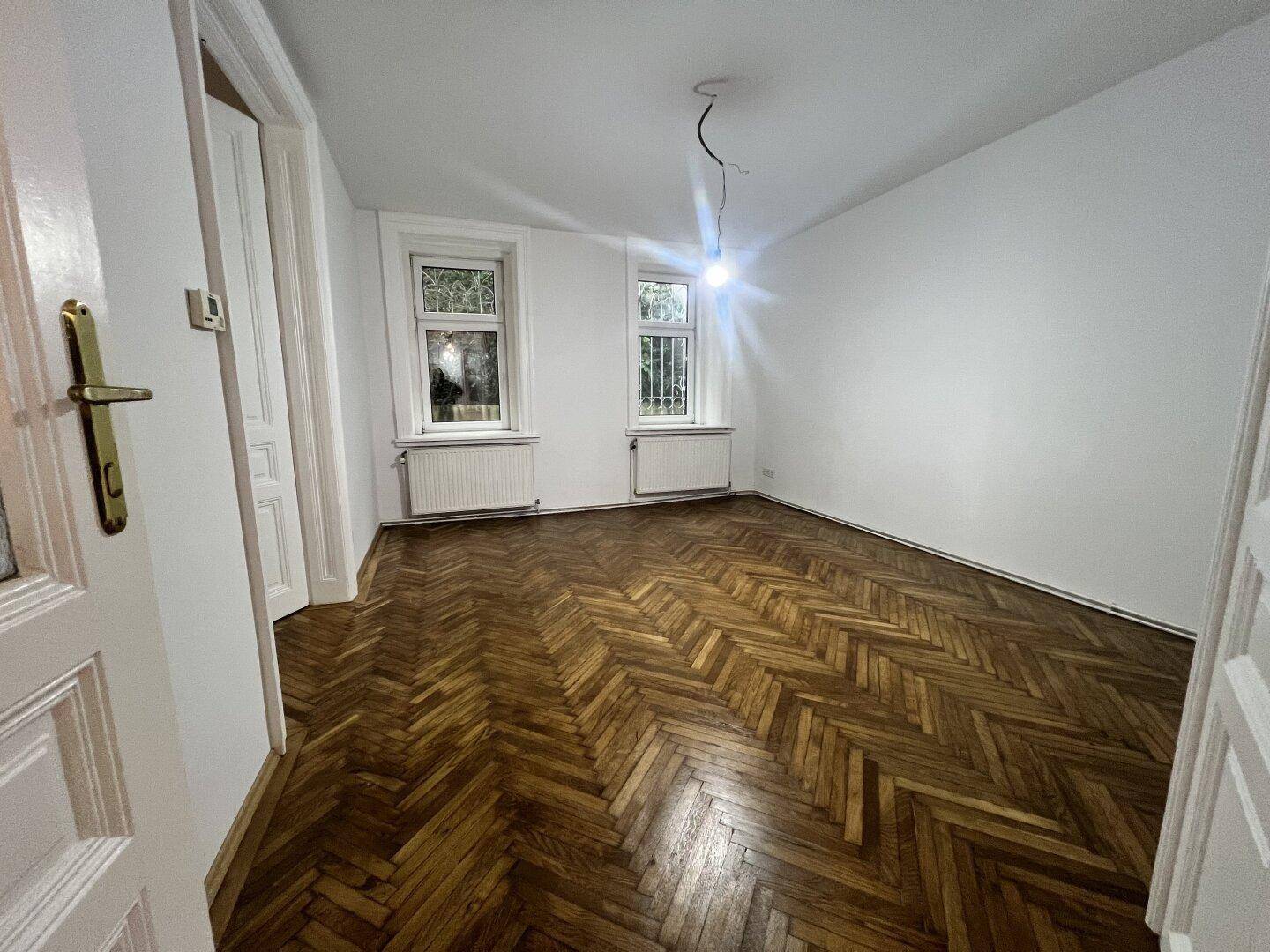 Geräumiges Zimmer mit klassischem Fischgrätparkett, weißen Wänden und viel Tageslicht.
