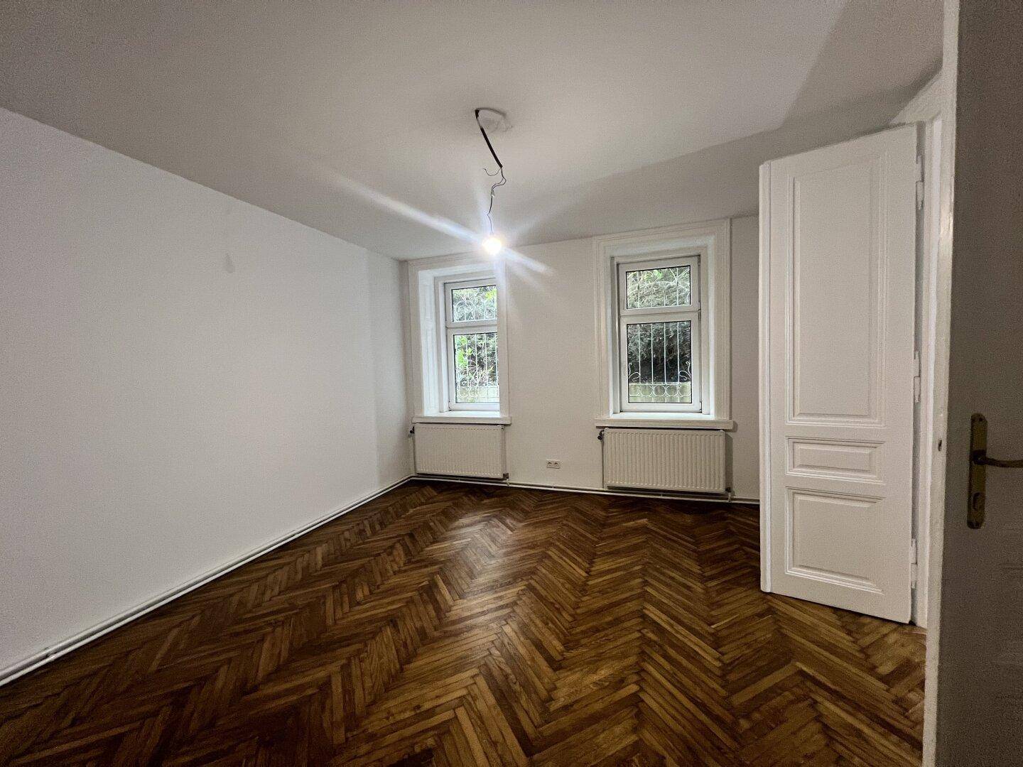 Helles Zimmer mit Fischgrätparkett, weißen Wänden und zwei Fenstern mit Blick ins Grüne.