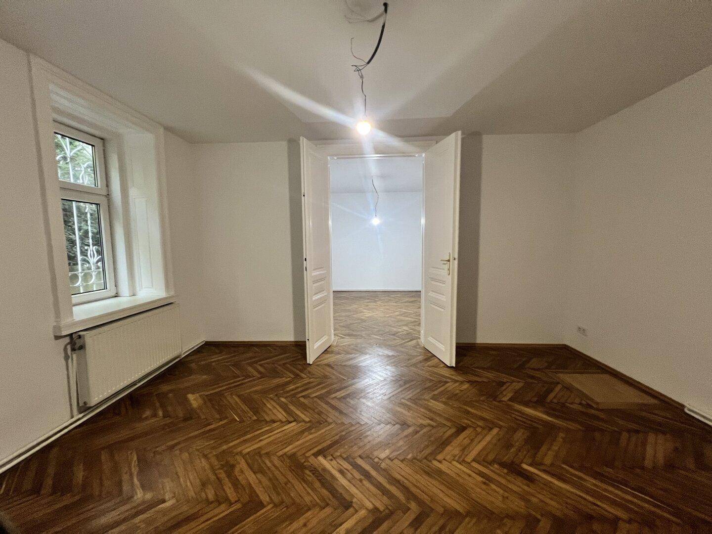 Heller Raum mit Fischgrätparkett und Doppeltüren, die zu einem weiteren Zimmer führen.