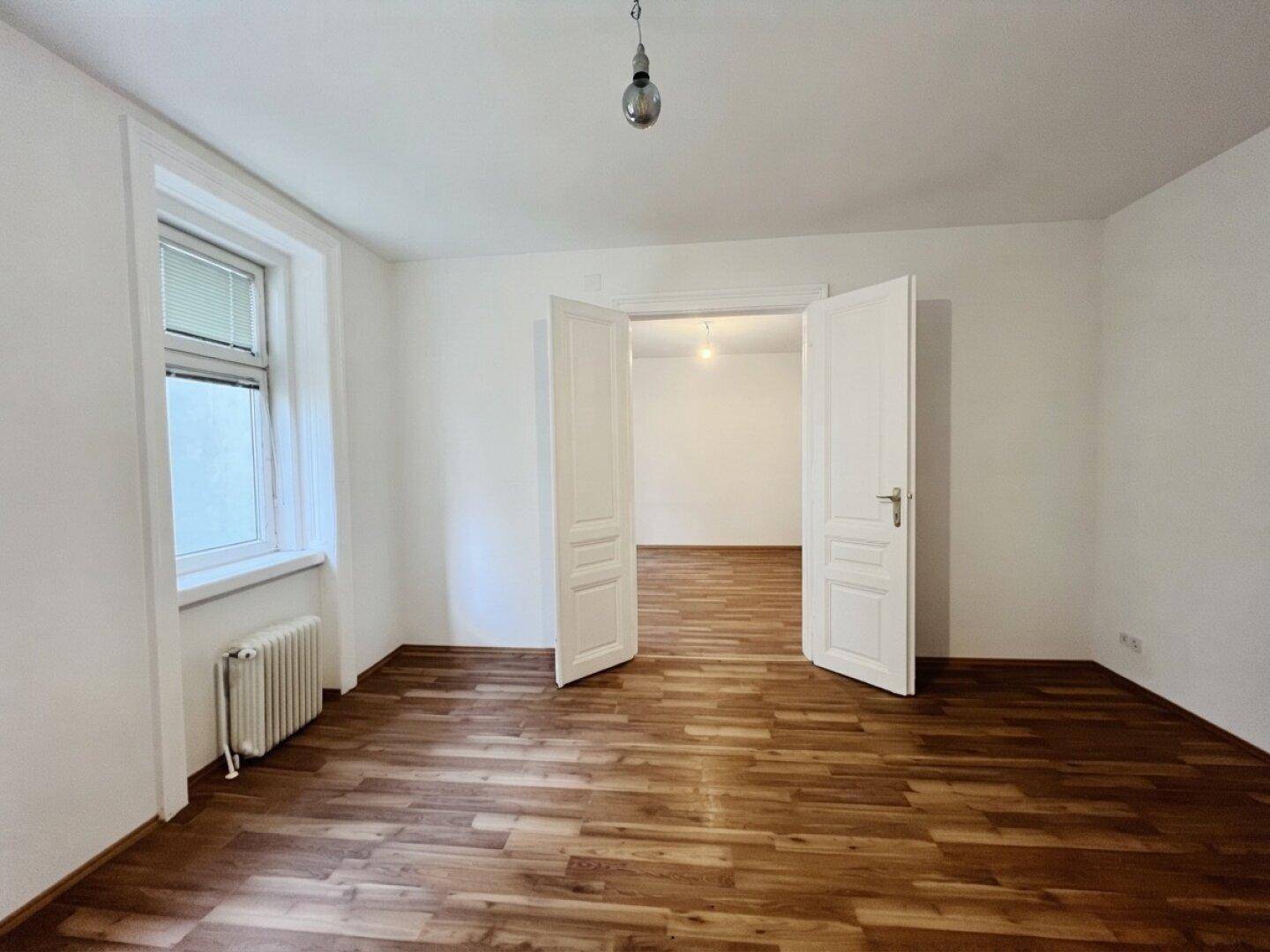 Lichtdurchflutetes Zimmer mit Parkettboden und Doppeltüren, das zu einem weiteren Raum führt.