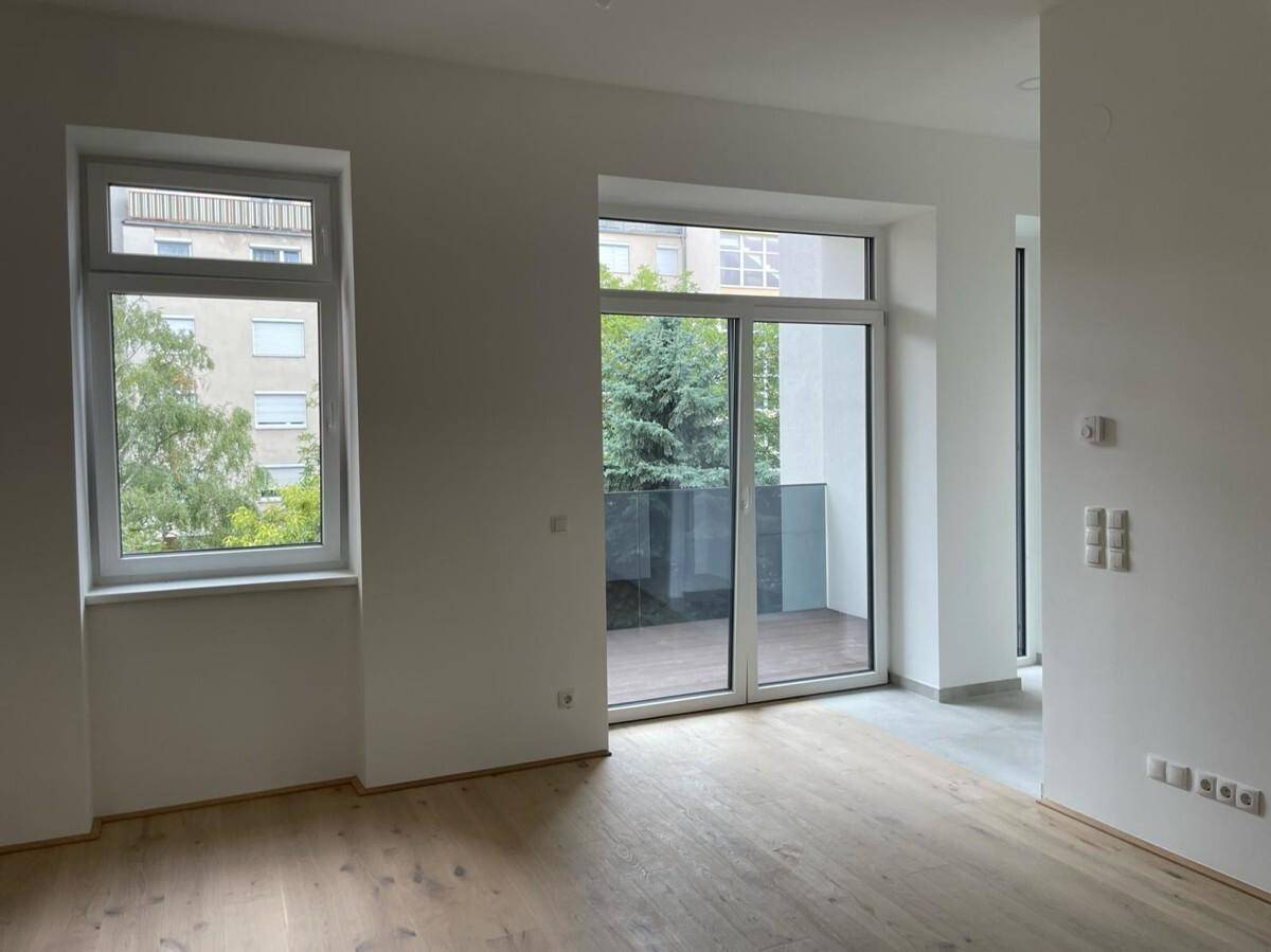 Heller, leerer Raum mit Holzboden und großem Fenster sowie Zugang zu einem Balkon.
