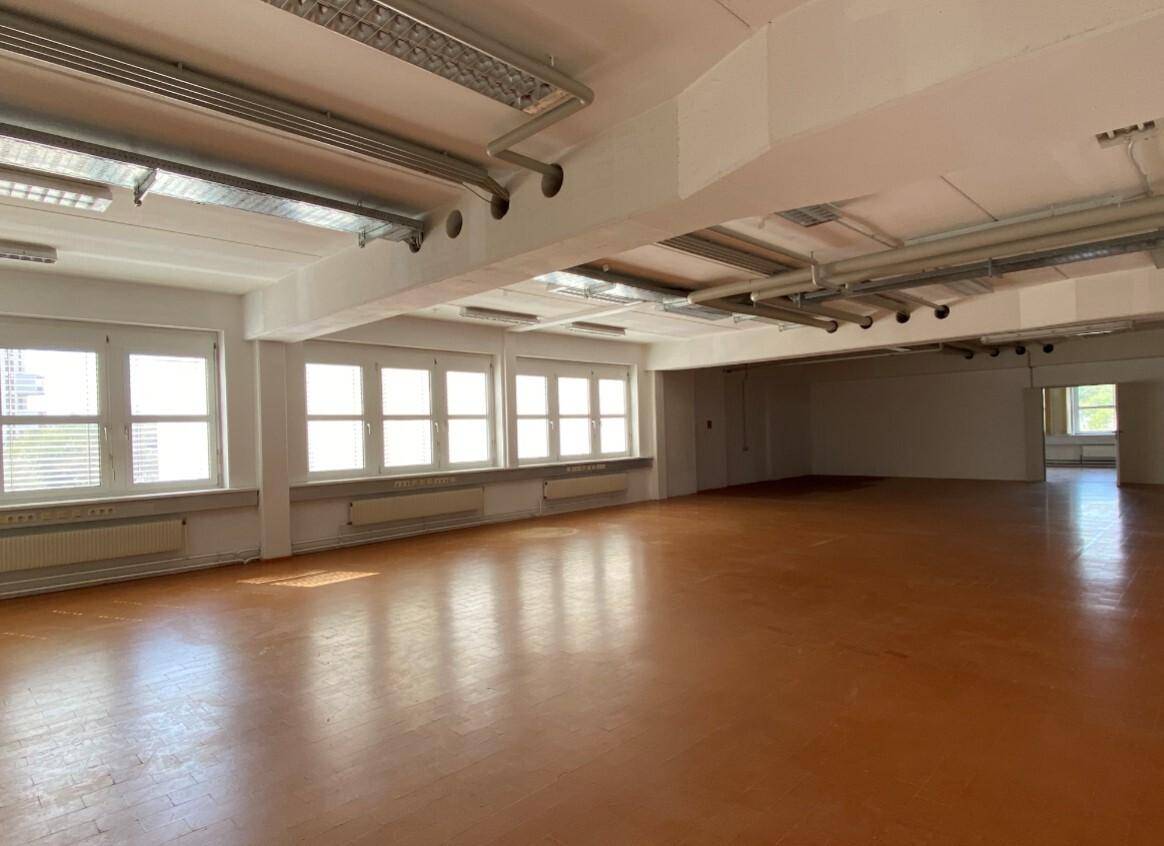 Heller Gewerberaum mit großen Fenstern, braunem Boden und sichtbaren Deckeninstallationen.