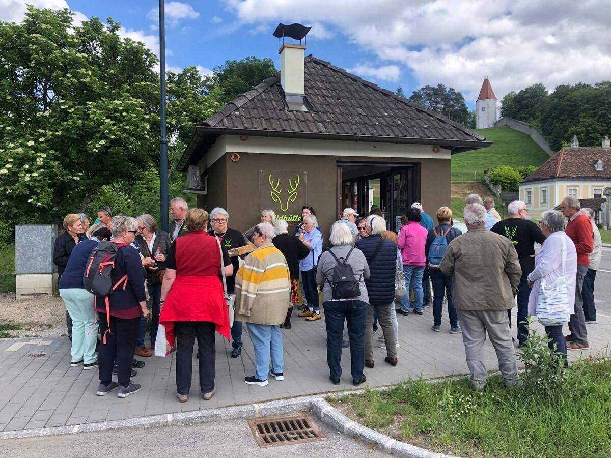 Eine Gruppe von Menschen versammelt sich vor dem kleinen Gebäude mit Ziegeldach an einem sonnigen Tag.