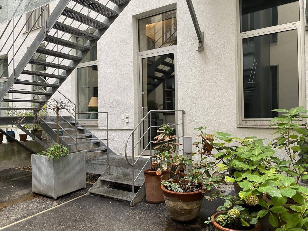 Hinterhofansicht mit moderner Metalltreppe, großen Fenstern und üppigen Pflanzen im Eingangsbereich.