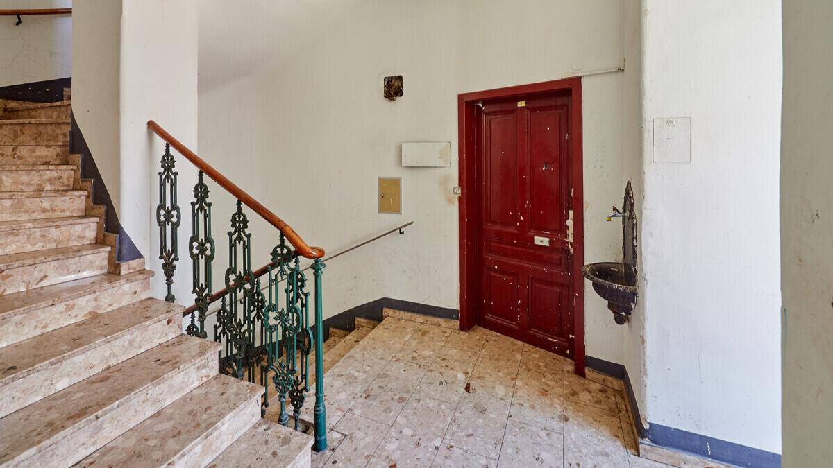 Historisches Treppenhaus mit Terrazzoboden, schmiedeeisernem Geländer und einer markanten roten Eingangstür.
